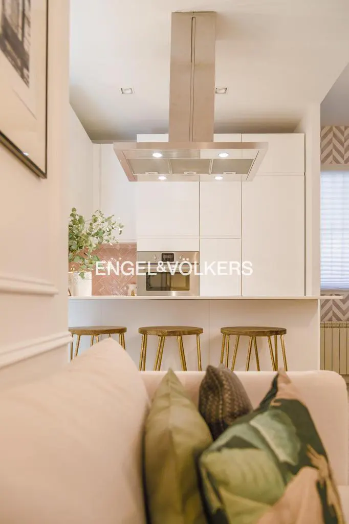 Interior view of a modern kitchen with white cabinets, a stainless steel range hood, and wooden stools at the counter. Pillows on a pink sofa are in the foreground.