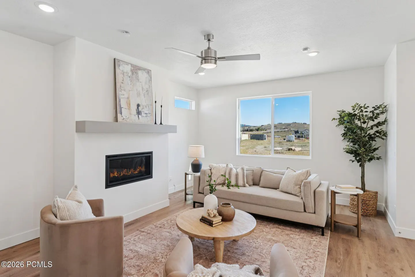 Modern living room with a fireplace, beige sofa, two armchairs, round wooden coffee table, and a view of a rural landscape.