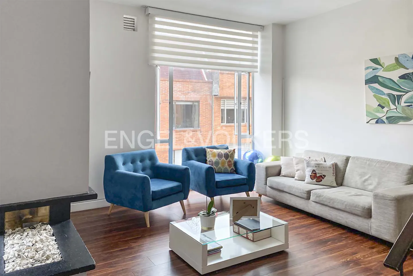 Bright living room with hardwood floors, blue armchairs, and a gray sofa. A white coffee table sits in the center, and a fireplace is on the left.