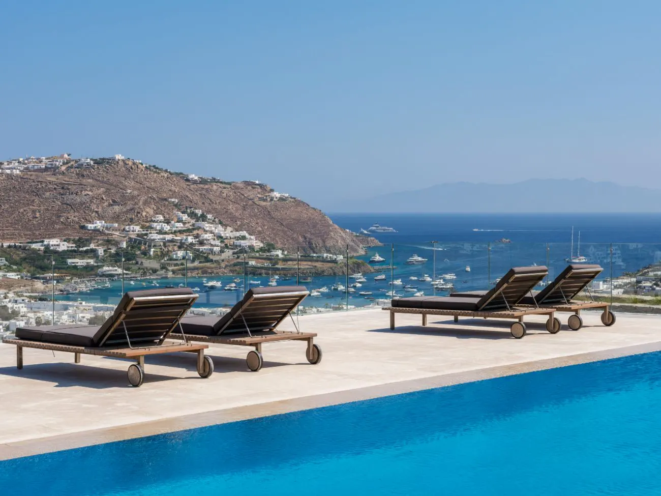 Luxury pool with lounge chairs overlooking a bay filled with boats and a hillside village under a clear blue sky.