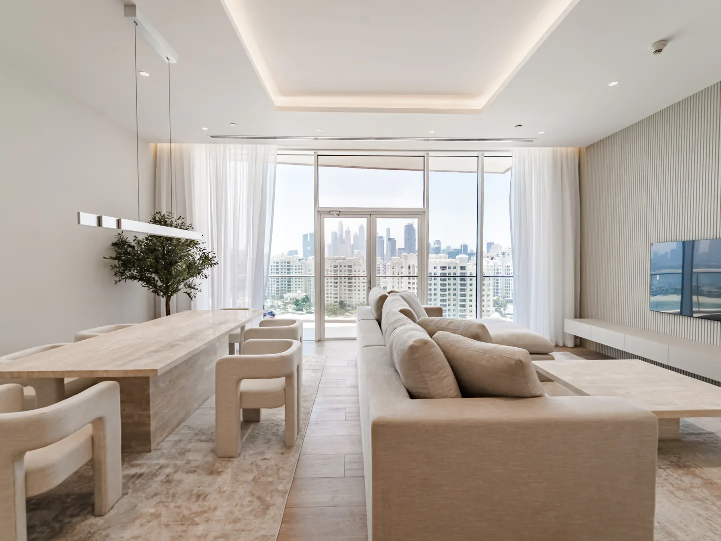 Bright, modern living room with beige sofa, table, and chairs. Large windows offer a city view. A flat-screen TV hangs on the wall.