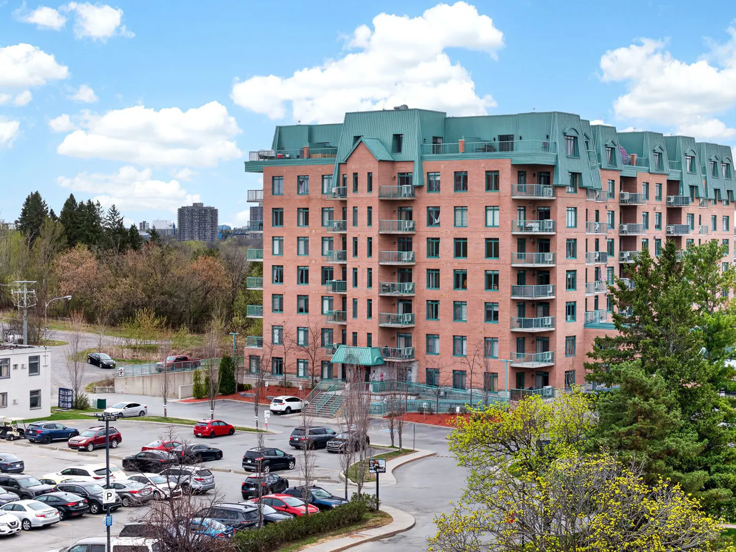 Exterior view of a brick apartment building with green roof and balconies, parking lot in front.