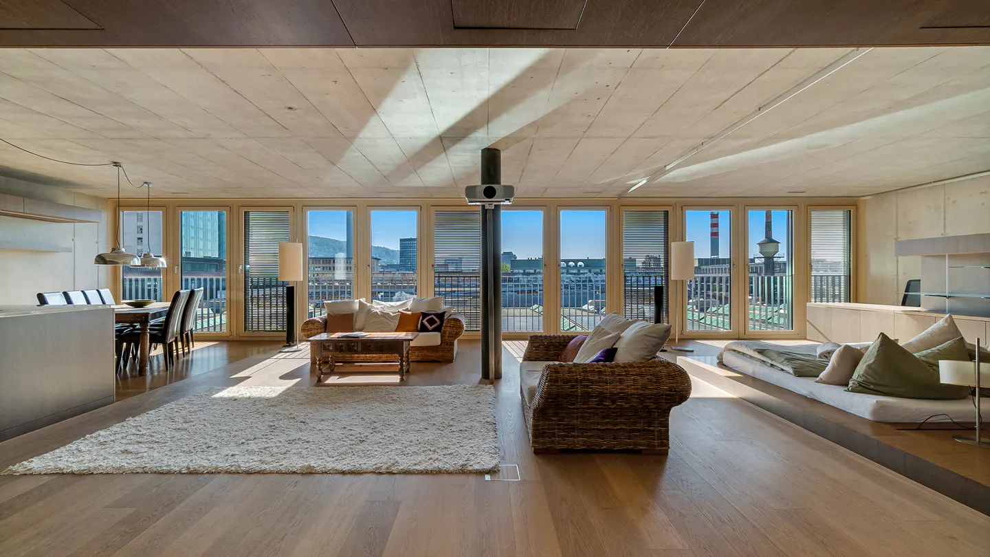 Bright, modern living room with wood floors, a white rug, and wicker sofas. Large windows offer a city view.