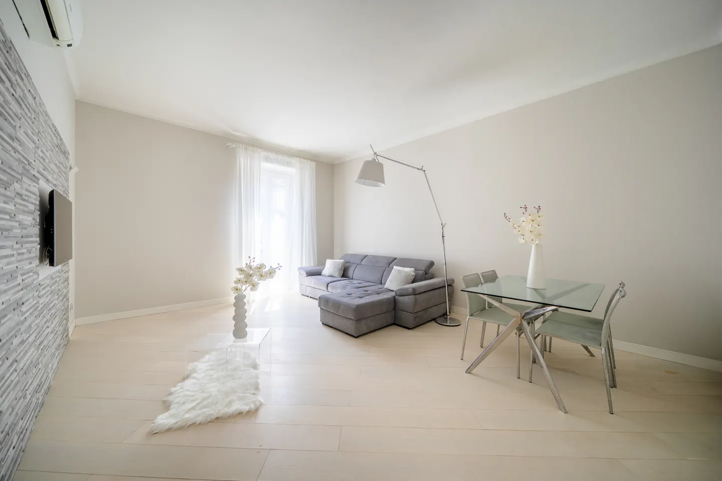 Bright living room with gray sectional sofa, glass table, and white walls. A TV hangs on a textured wall.