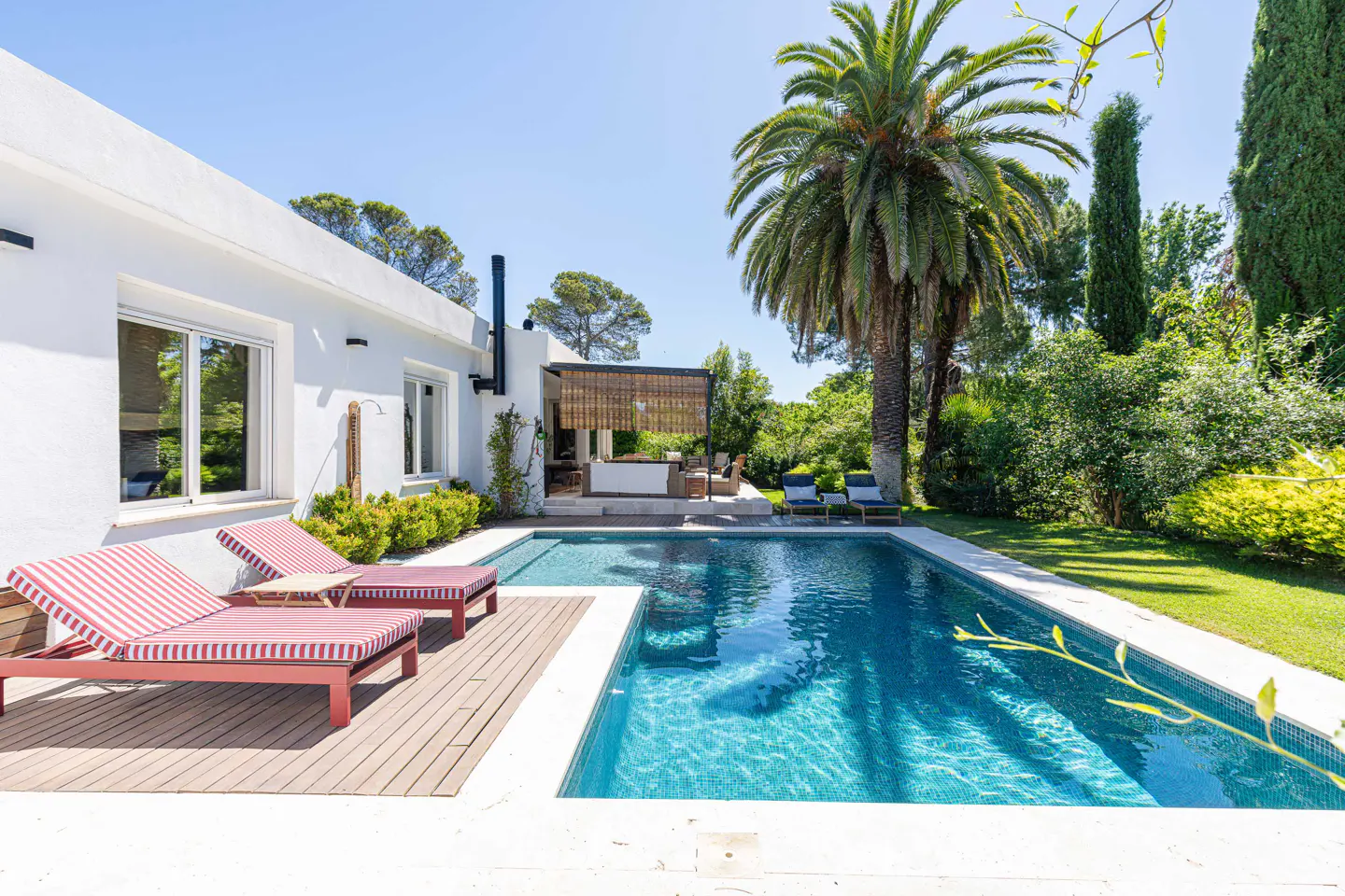 A white house with a swimming pool, red-striped lounge chairs on a wooden deck, a palm tree, and a lush green garden.