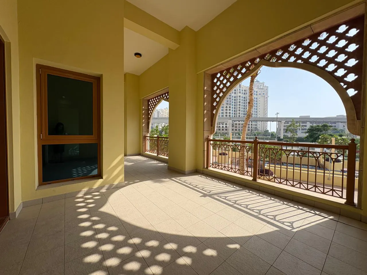 A sunny balcony with tan tile flooring, decorative archways, and a view of a city skyline.