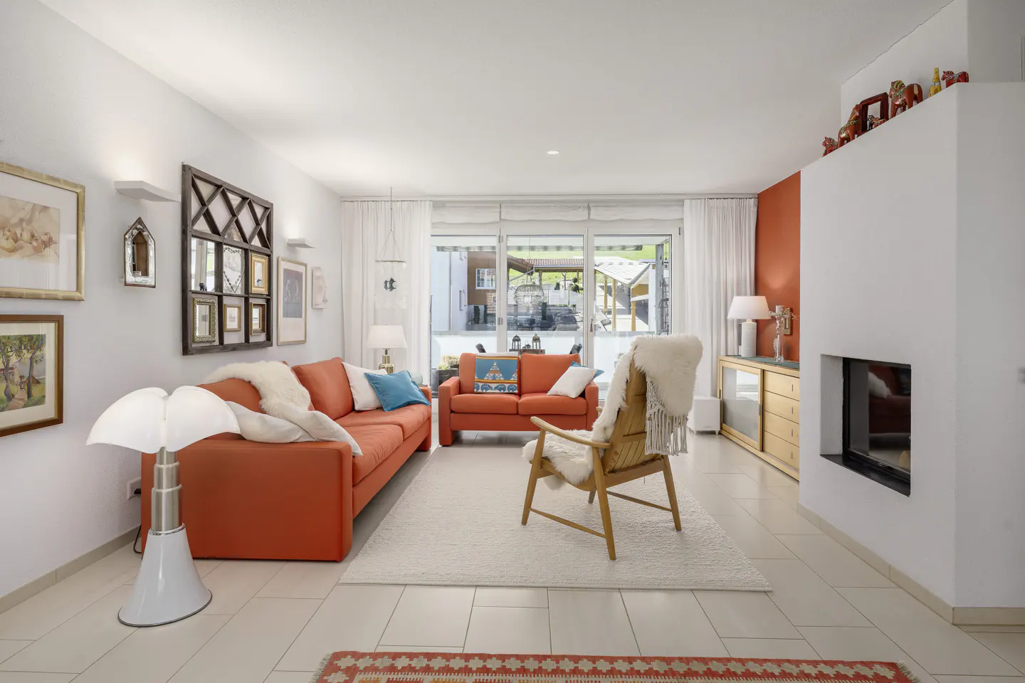 Bright living room with orange sofas, a wooden chair with a sheepskin throw, and a white fireplace. Large windows let in natural light.