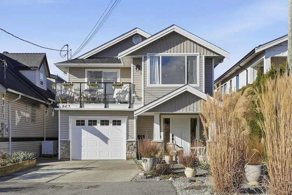 Two-story house with gray siding, white trim, and a balcony with chairs and a table above the garage. Tall ornamental grass in front.