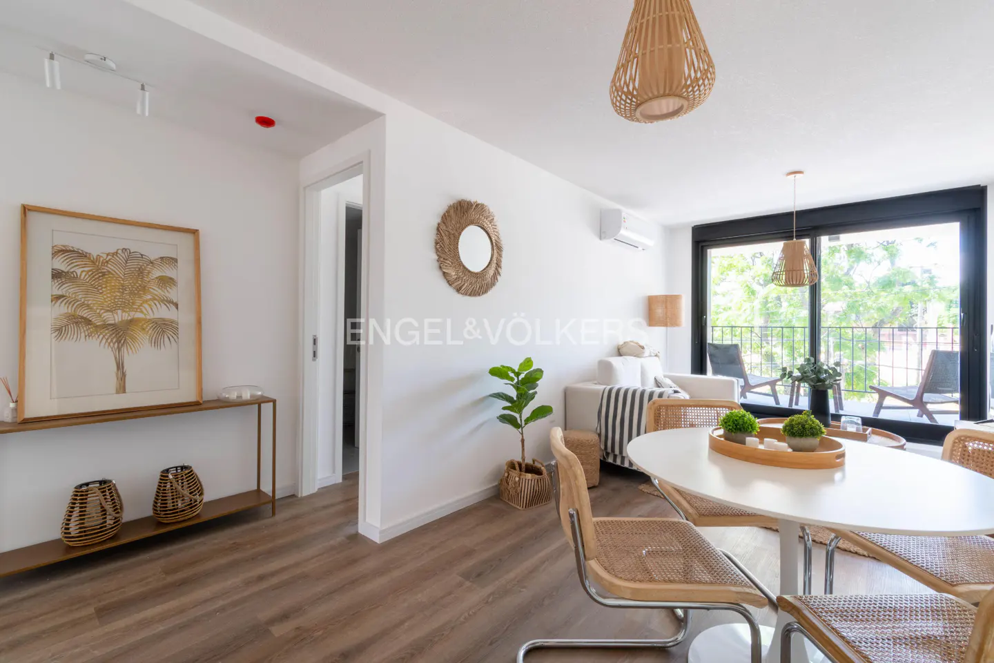 Bright living room with white walls, wood floors, and a round dining table with wicker chairs. A palm tree print hangs above a console table. Balcony view.