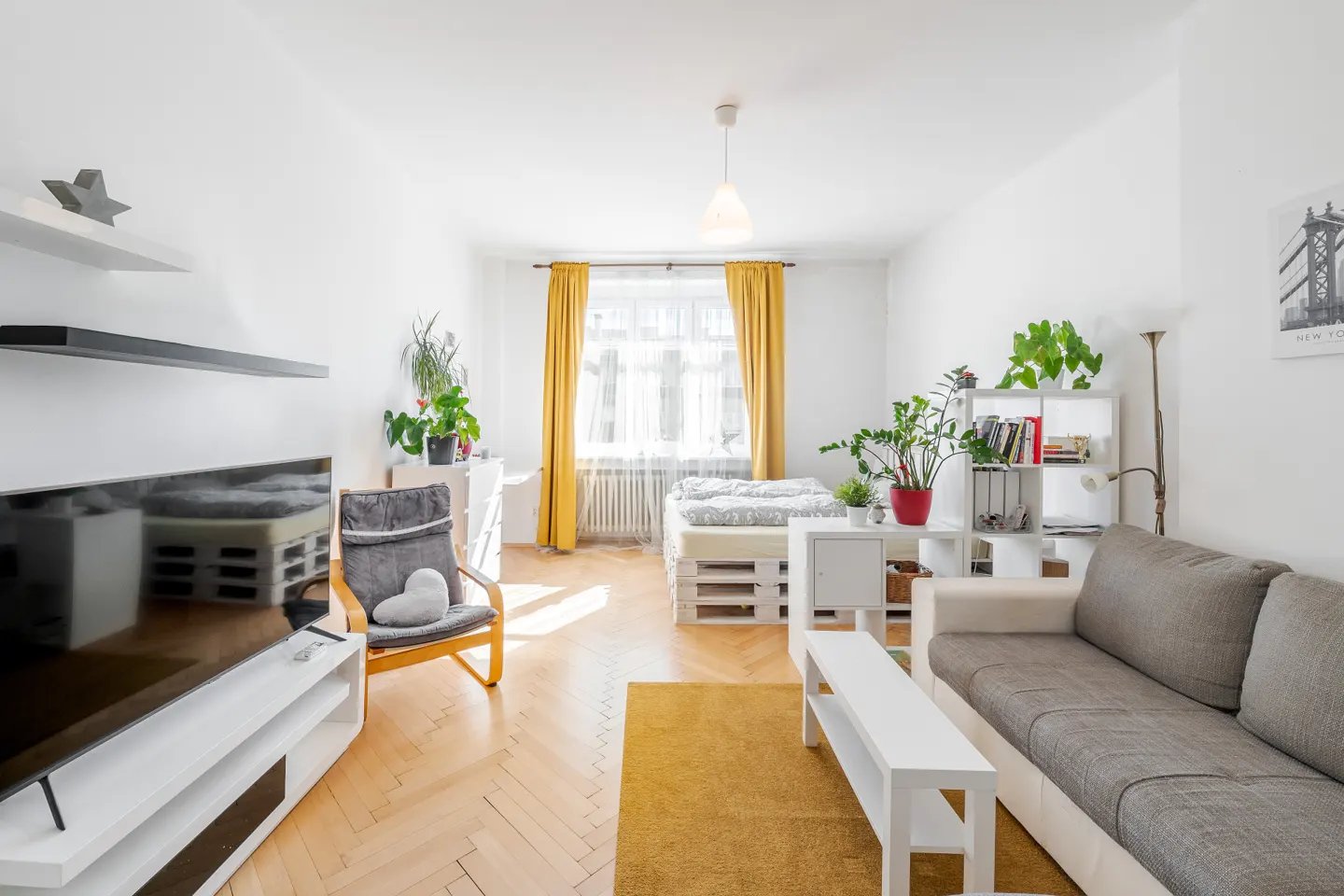 Bright studio apartment with a grey sofa, yellow rug, pallet bed, and large window with yellow curtains.