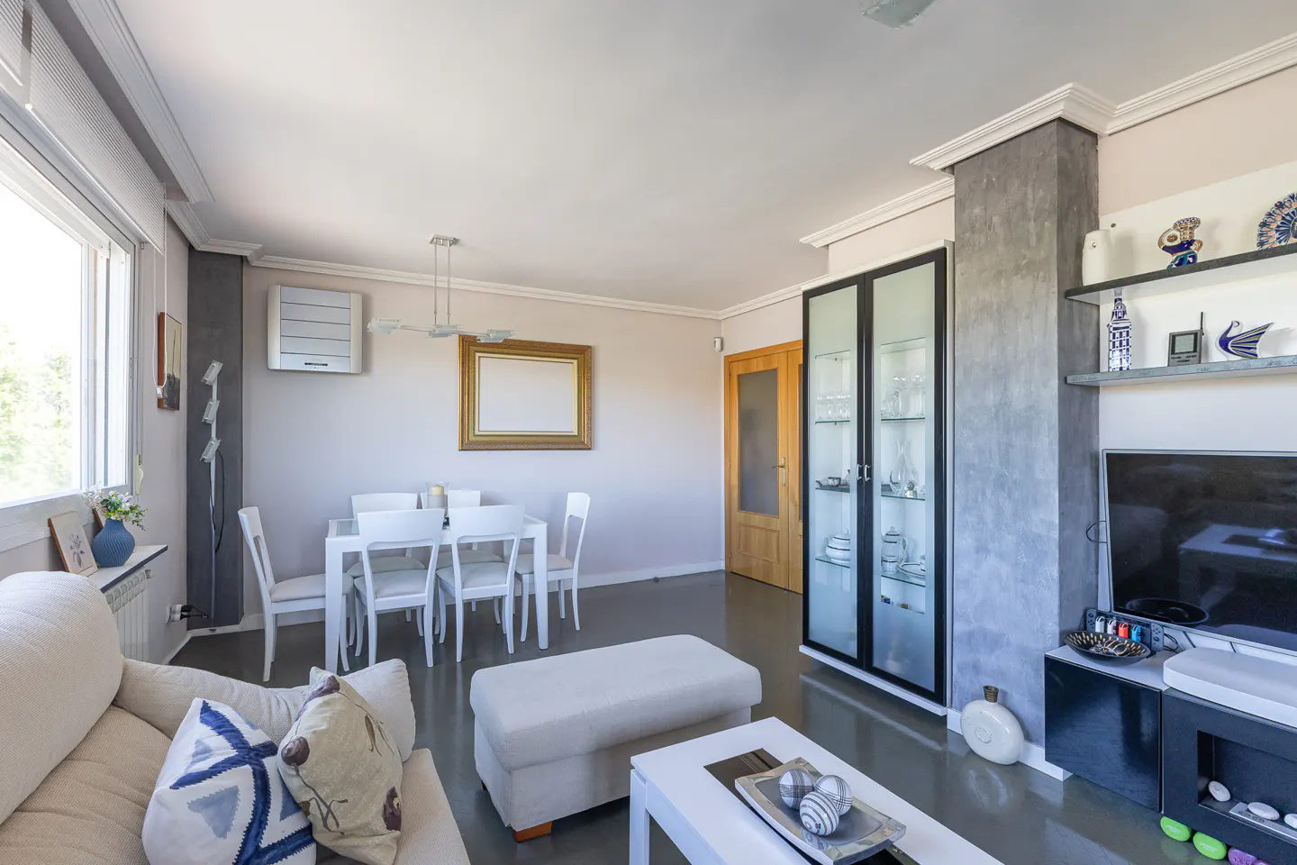 Modern living room with a white sofa, dining area, glass display cabinet, and a television on a grey feature wall.