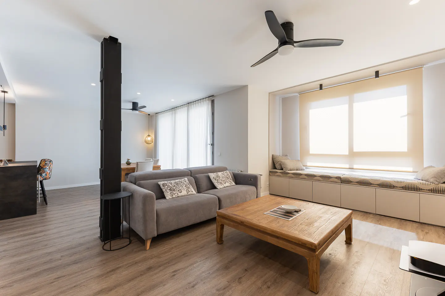 Living room with gray sofa, wooden coffee table, and window seat. A black support beam and ceiling fan add modern touches. Light wood floors.