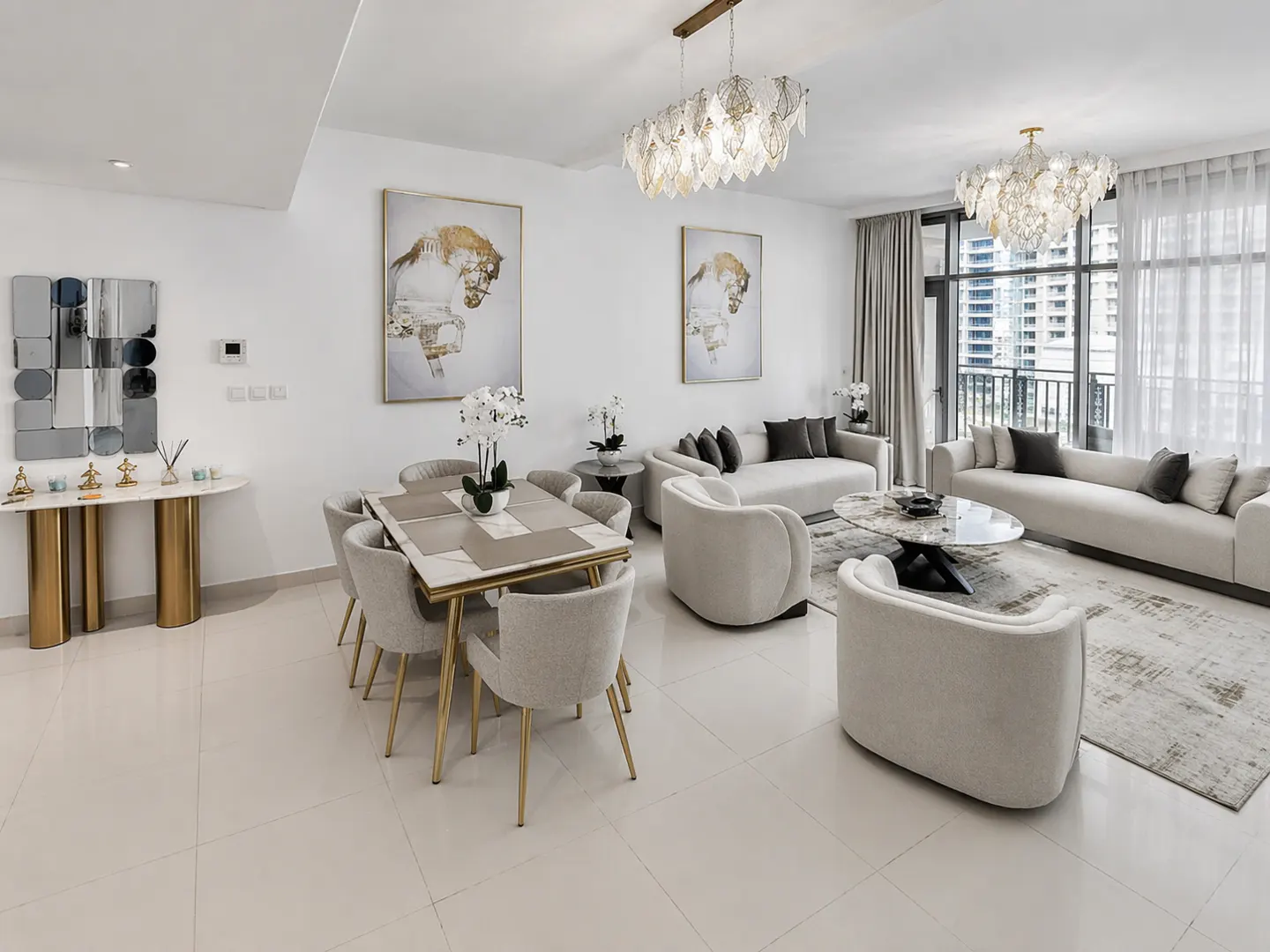 Bright, modern living and dining area with white walls, tile floors, and neutral furniture. Two chandeliers hang above. Large windows show a city view.