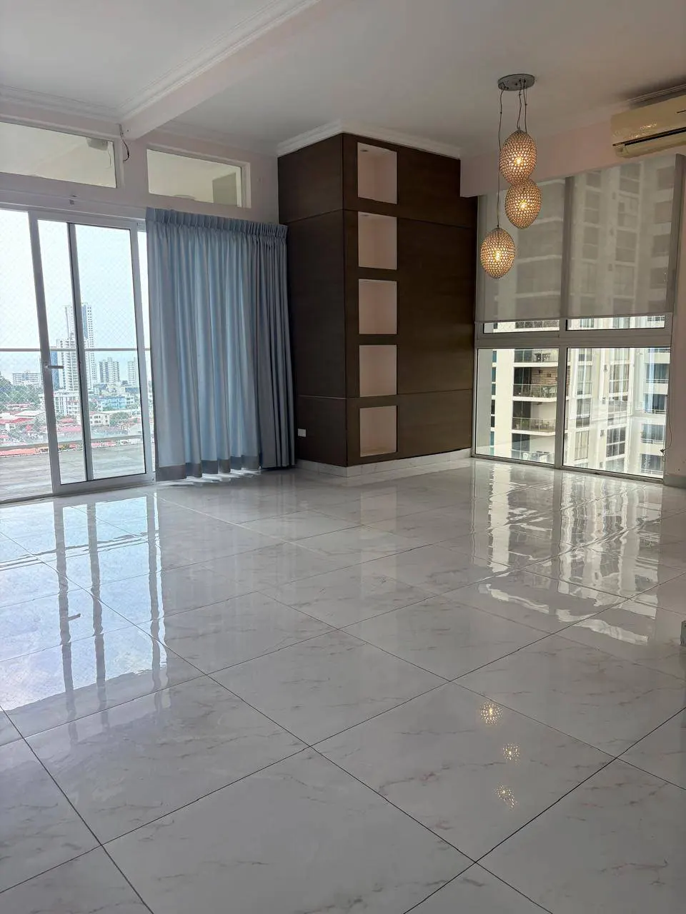 Bright, empty room with marble floors, floor-to-ceiling windows, and city views. Blue curtains and a brown built-in cabinet are visible.