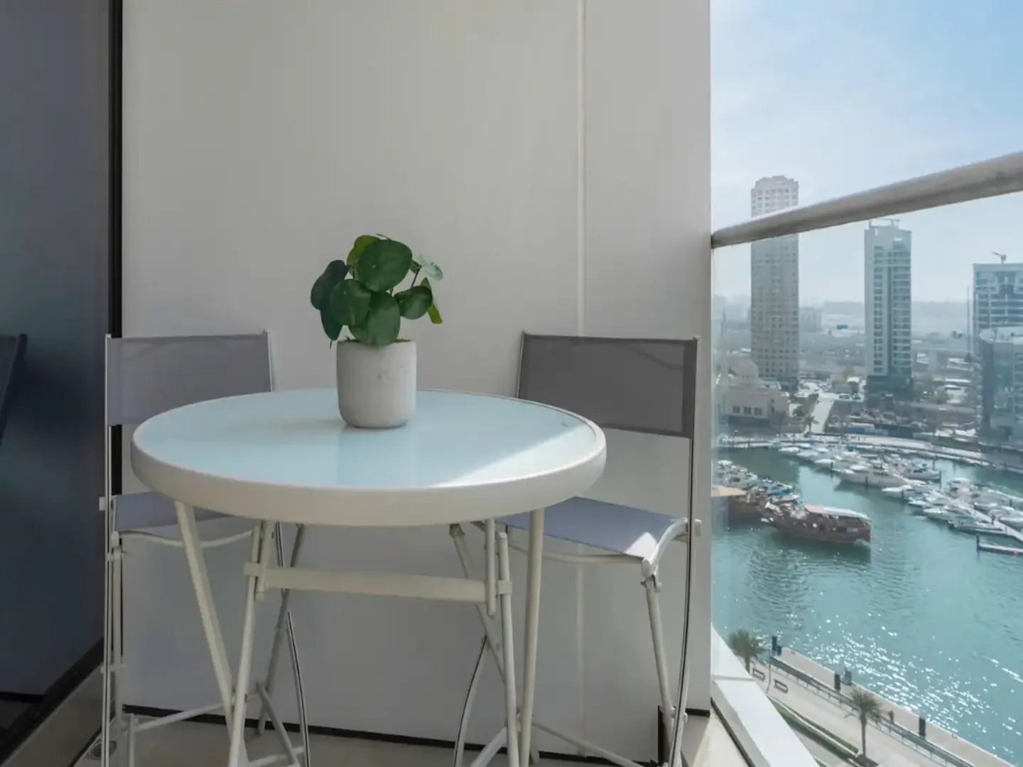 Small white balcony table with a potted plant and two chairs, overlooking a marina with boats and city skyscrapers.