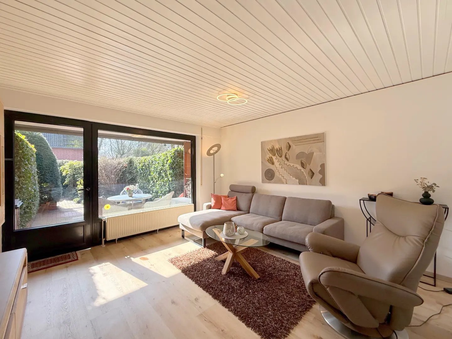 Living room with a gray sectional sofa, brown recliner, and glass coffee table on a brown rug. Large windows overlook a green garden.