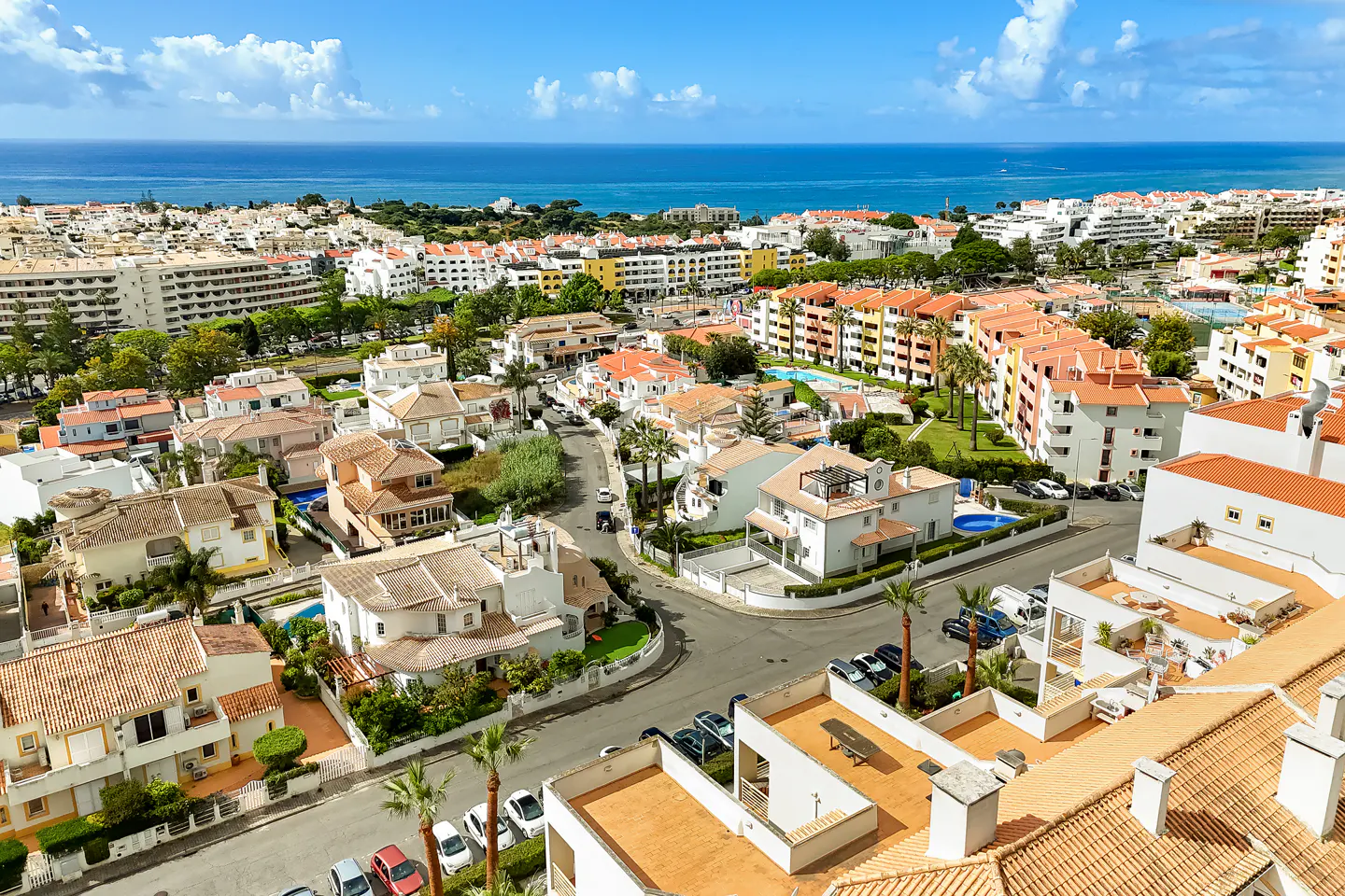 Aerial view of a coastal town with white buildings, orange roofs, palm trees, and the blue ocean in the background.