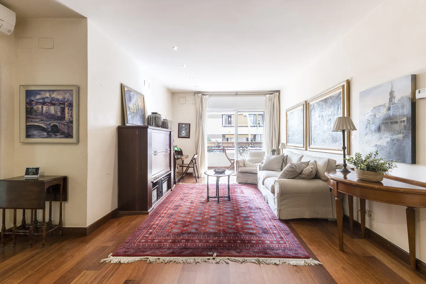 Living room with hardwood floors, a red rug, a white sofa, and paintings on the walls. Balcony in the background.