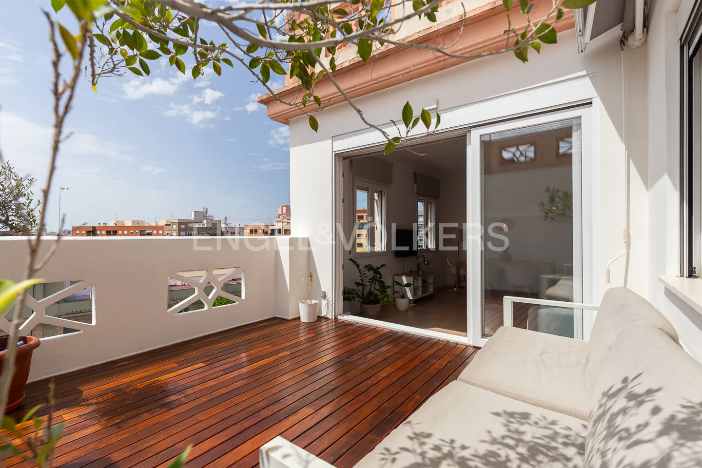 A bright, sunny balcony with a wood floor, white railing, and a beige sofa. Sliding glass doors lead to an interior living space.