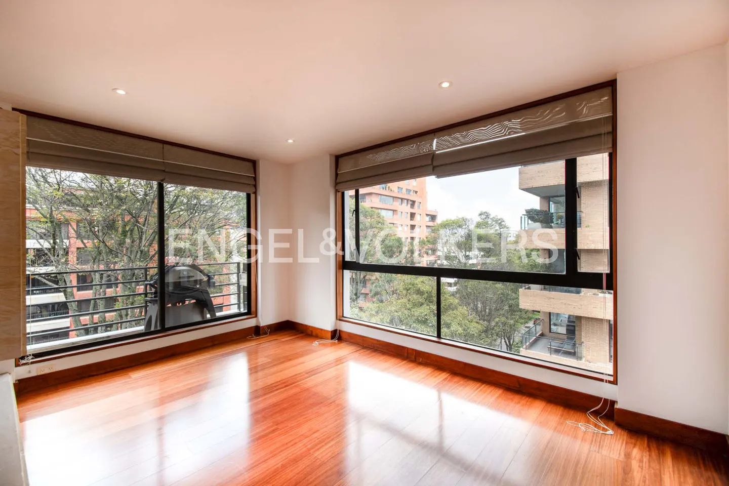Bright, empty room with hardwood floors and large windows. Outside, trees and buildings are visible. The windows have brown frames and shades.