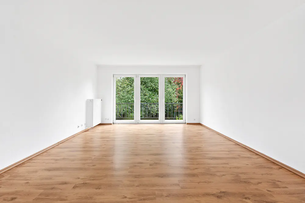 Empty room with wood floors, white walls, and a balcony with trees visible through the glass doors.