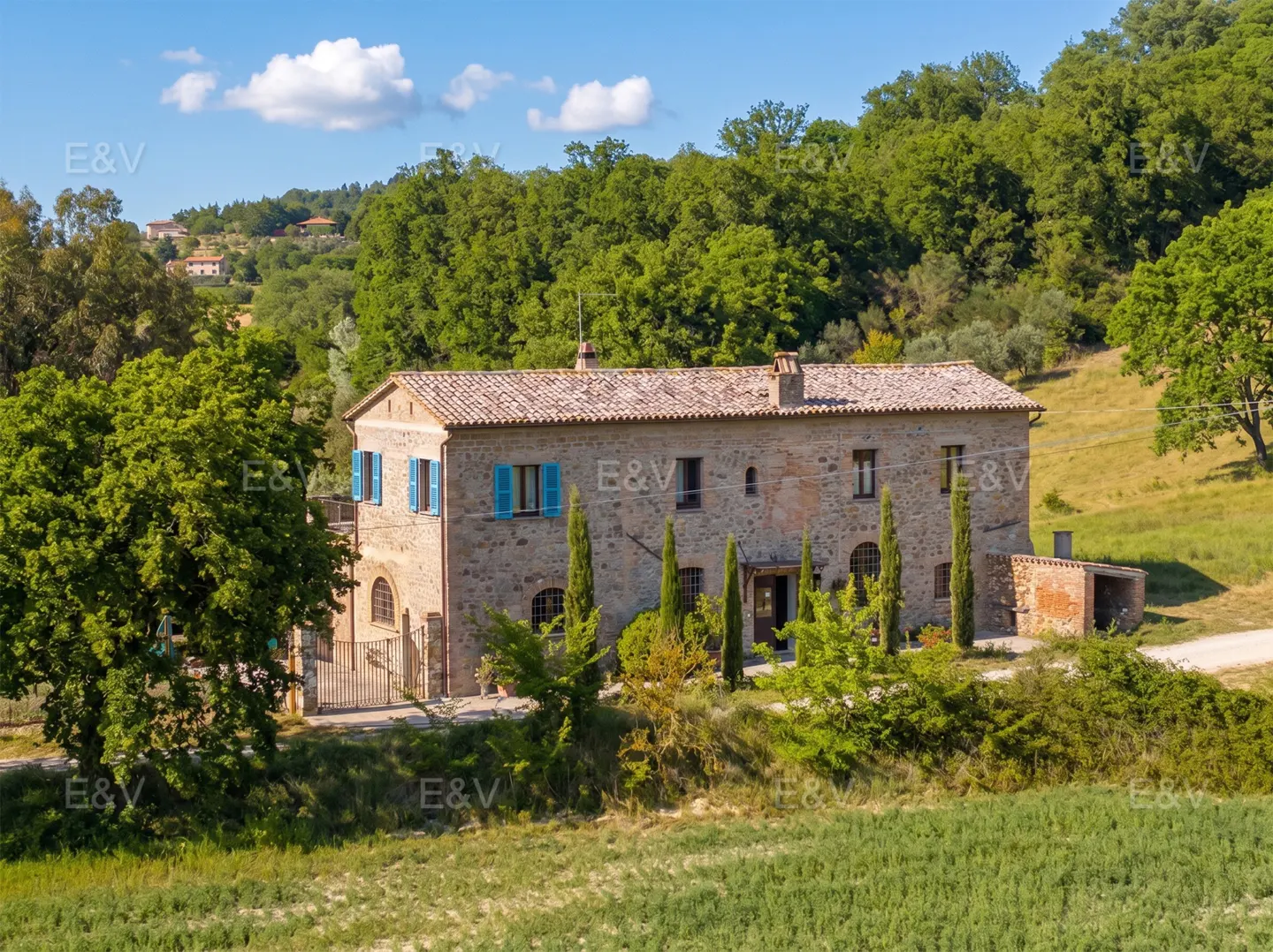 Exterior view of a two-story stone house with blue shutters, surrounded by green trees and fields under a blue sky.