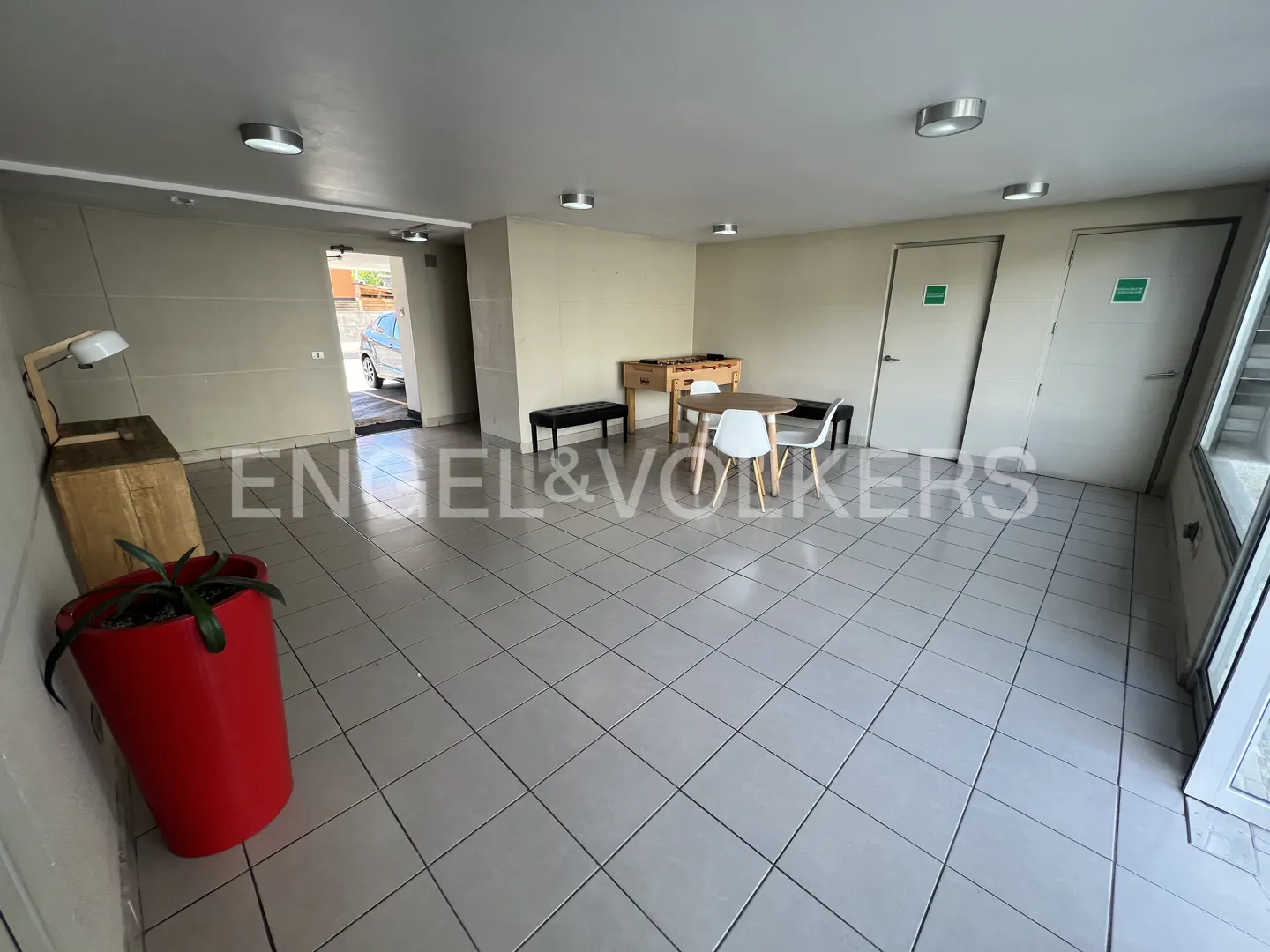 A common area with a foosball table, round table with chairs, and a red potted plant. The floor is tiled.