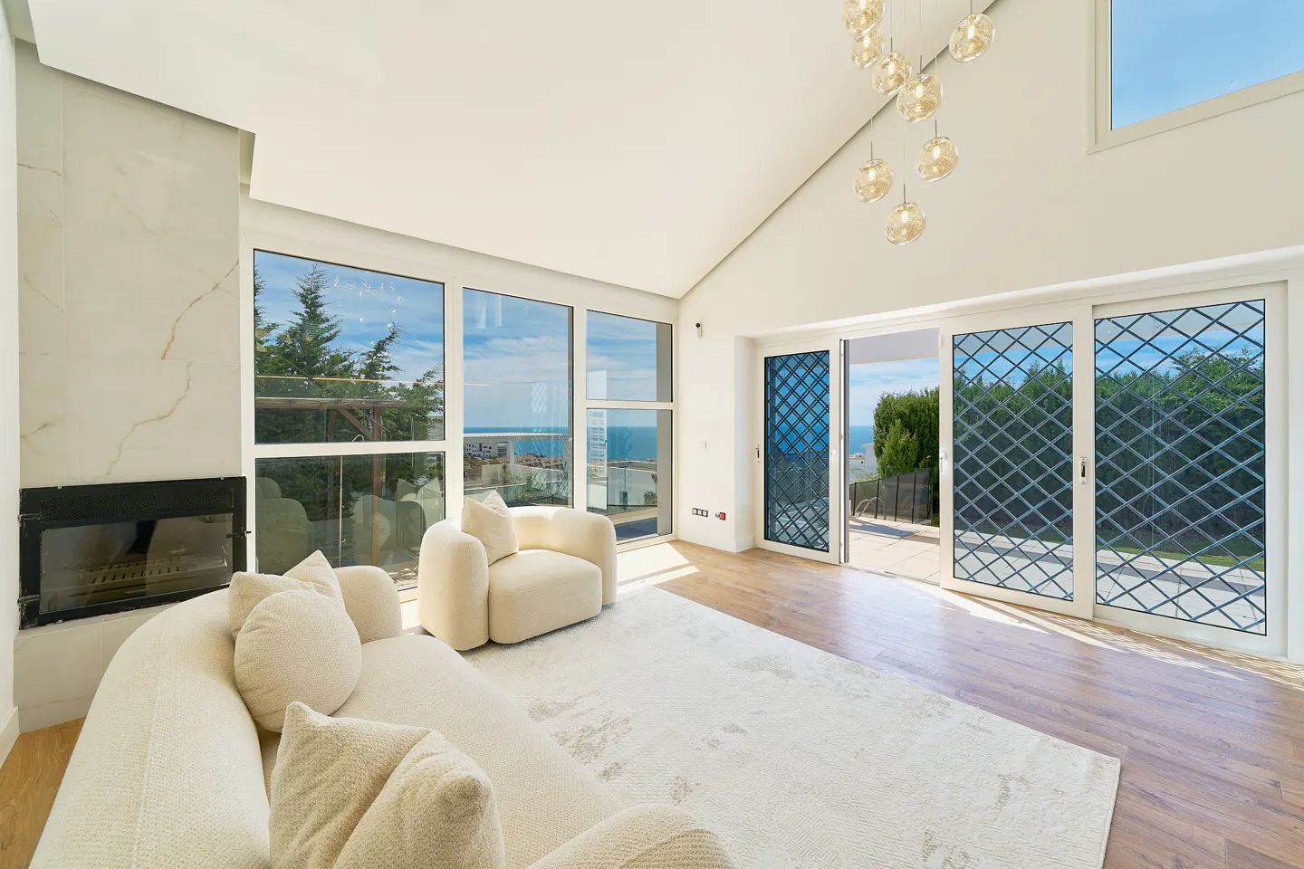 Bright living room with white sofa, chair, and rug. Large windows offer ocean views. A fireplace and modern chandelier add elegance.