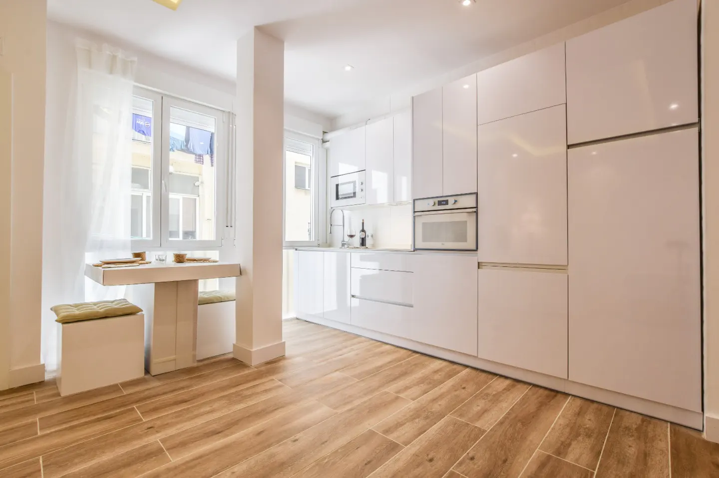 Modern kitchen with white glossy cabinets, built-in appliances, wood-look tile floors, and a small dining nook by the window.