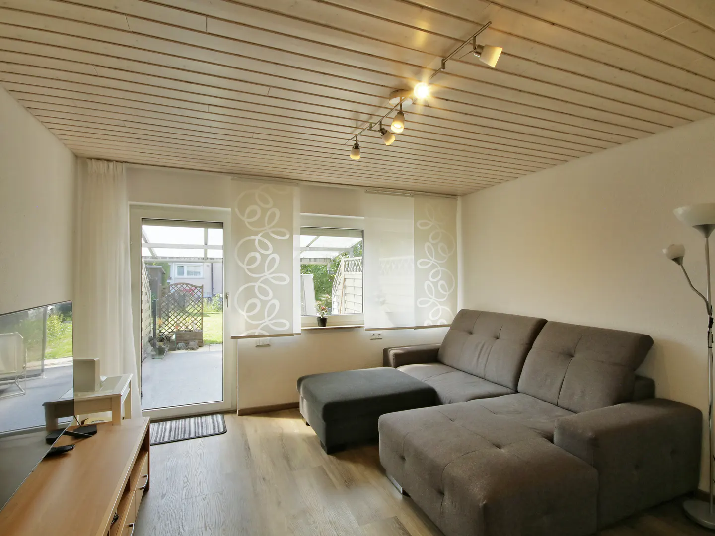 Living room with a gray sectional sofa, wood floors, and a door leading to a patio. The ceiling is wood paneled with track lighting.