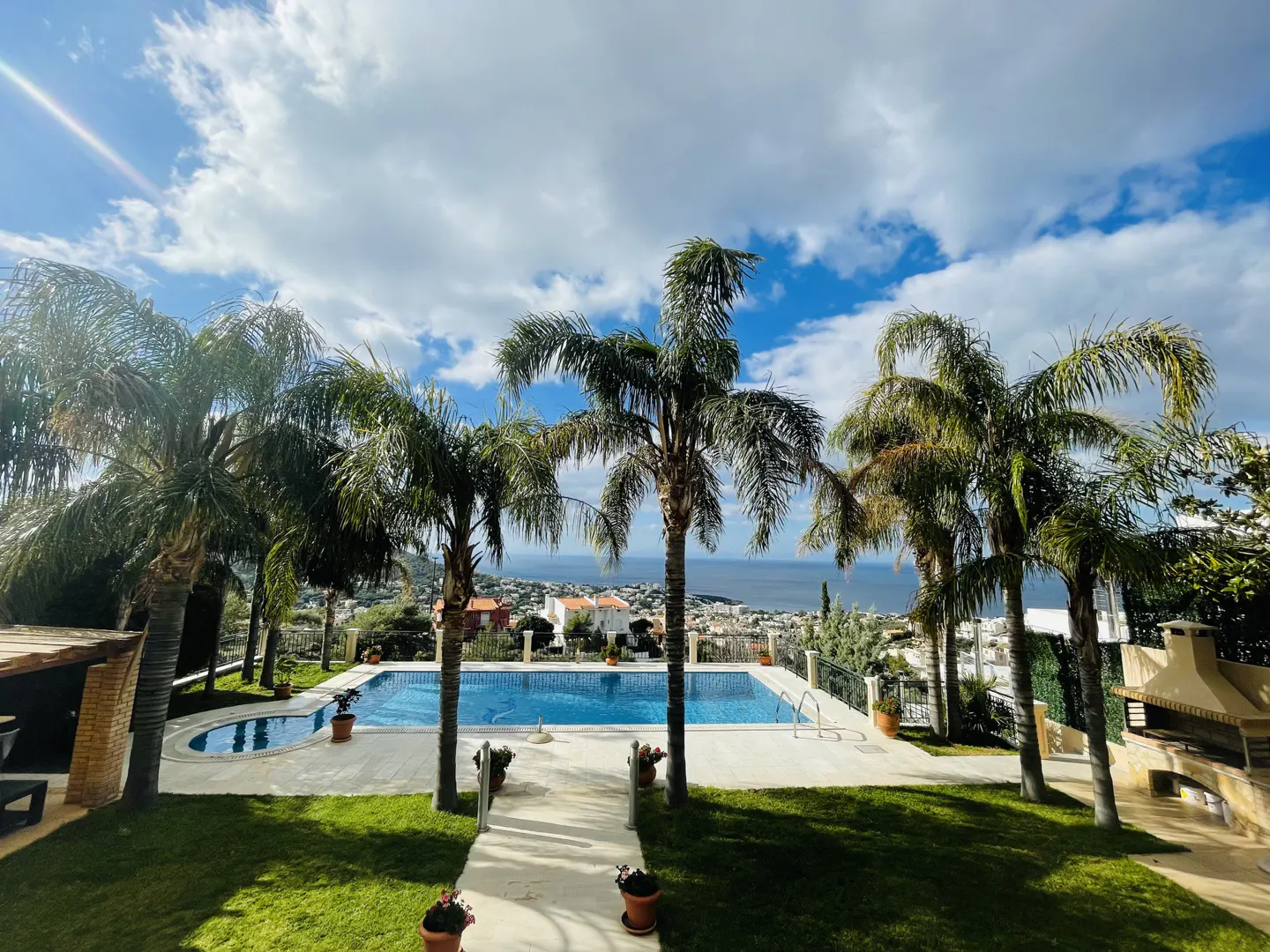 Outdoor view of a blue swimming pool surrounded by palm trees and green grass, with a city and ocean in the background.