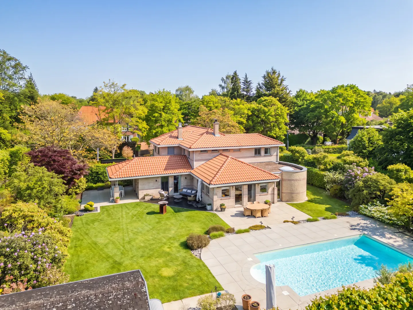 Aerial view of a large, tan house with a red tile roof, a green lawn, a patio, and a blue swimming pool surrounded by lush green trees.