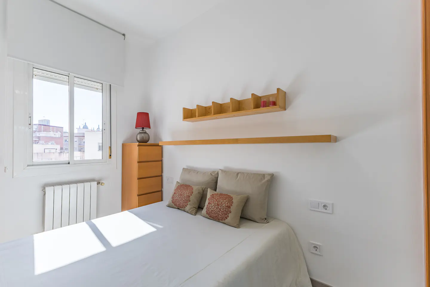 Bright bedroom with white walls, a bed with beige pillows, and wooden shelves. A window lets in sunlight, illuminating the room.