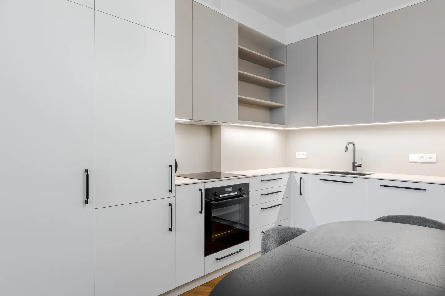 Modern kitchen with white and grey cabinets, built-in oven, under-cabinet lighting, and a grey dining table in the foreground.