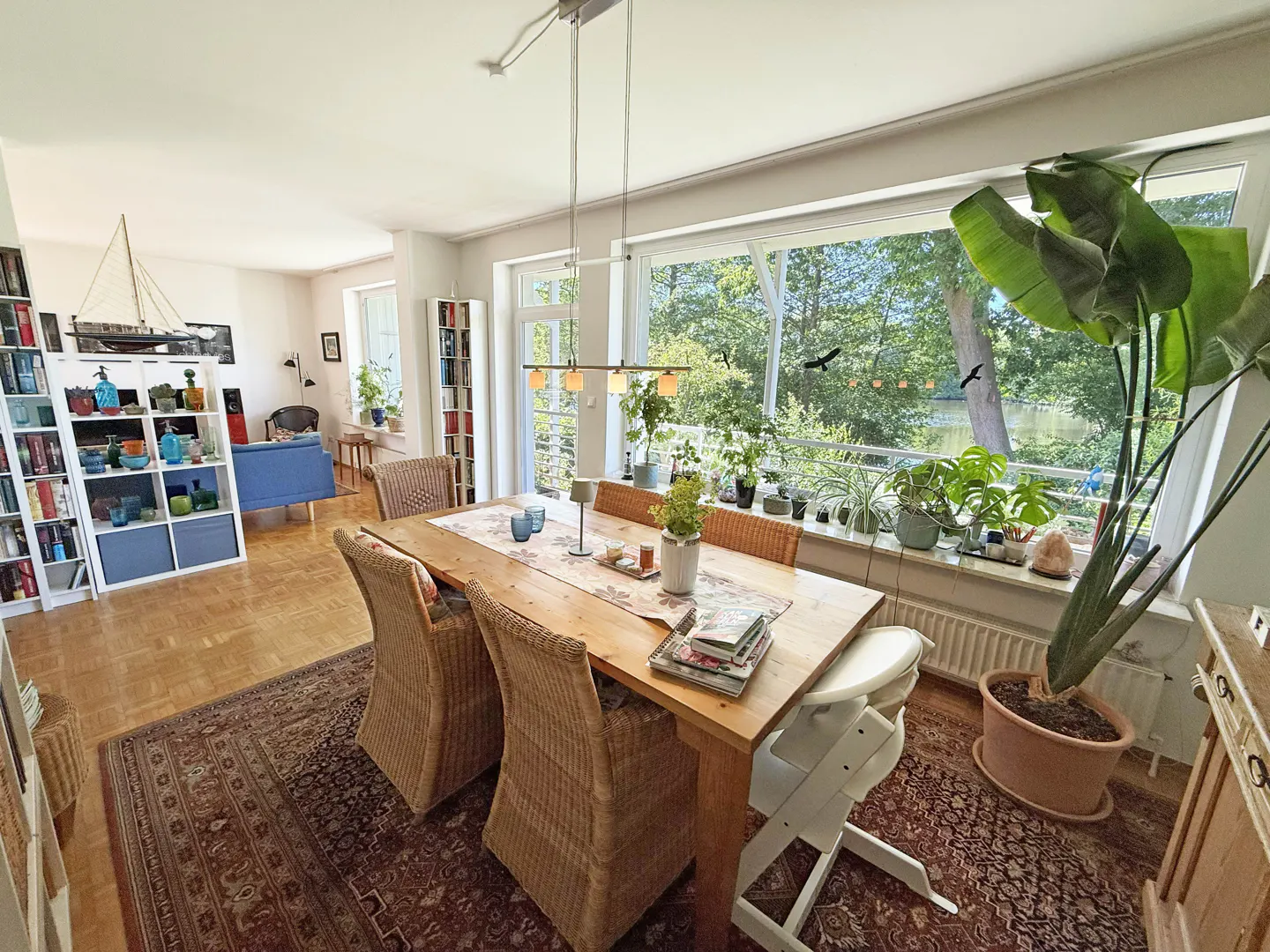Bright dining room with a wooden table, wicker chairs, large windows overlooking trees, and a large potted plant.