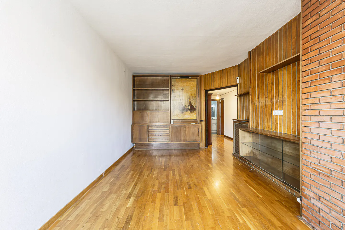 A bright room with hardwood floors, a white wall, and a wooden cabinet with shelves and drawers. A painting hangs next to the cabinet.