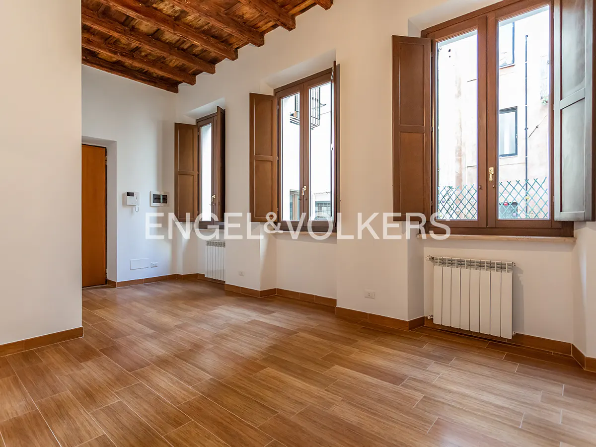 An empty room with wood floors, white walls, and a wood-beamed ceiling. Three windows with brown shutters let in natural light. Radiators are under each window.