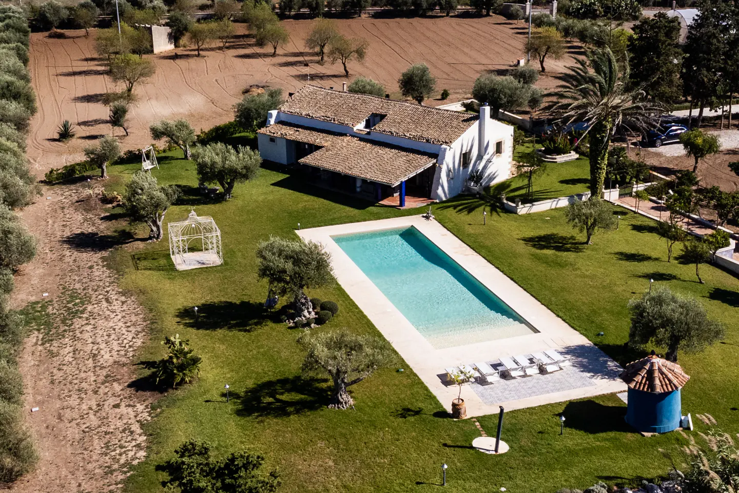 Aerial view of a white house with a brown tile roof, a blue-tiled porch, and a rectangular pool surrounded by green grass and trees.