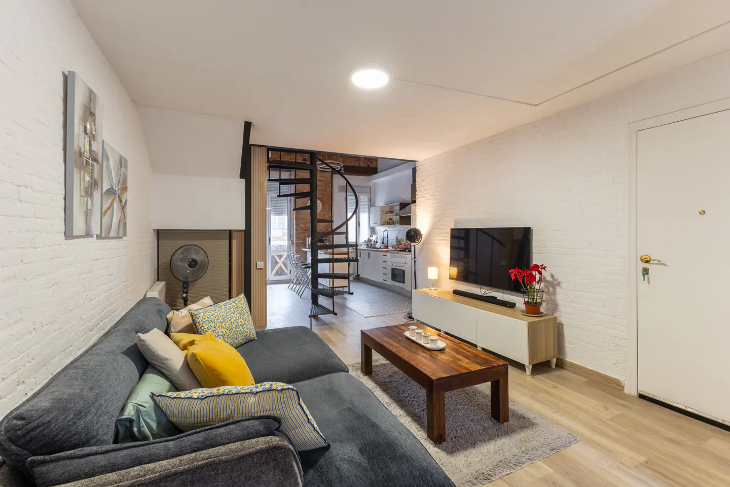Living room with gray sectional sofa, wood coffee table, TV, and spiral staircase leading to kitchen. White brick walls.