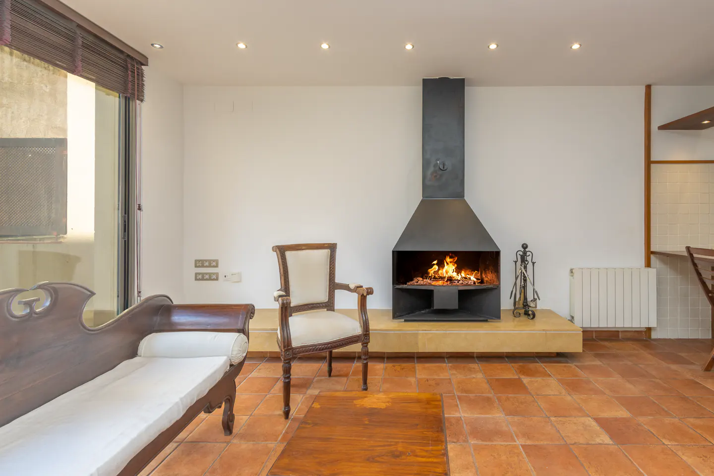Living room with a black fireplace, a wooden sofa, a chair, and a wooden table on a terracotta tile floor.