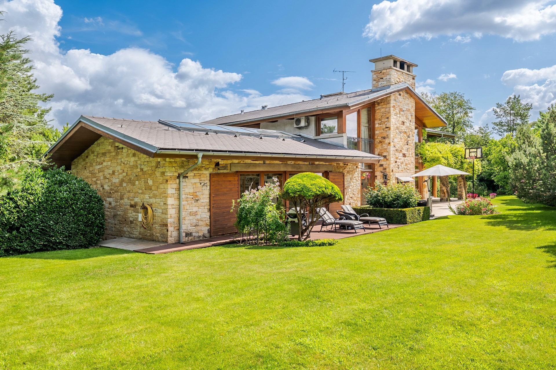 Modern stone house with a large lawn, patio furniture, and solar panels under a blue sky with fluffy white clouds. [AI-generated text]