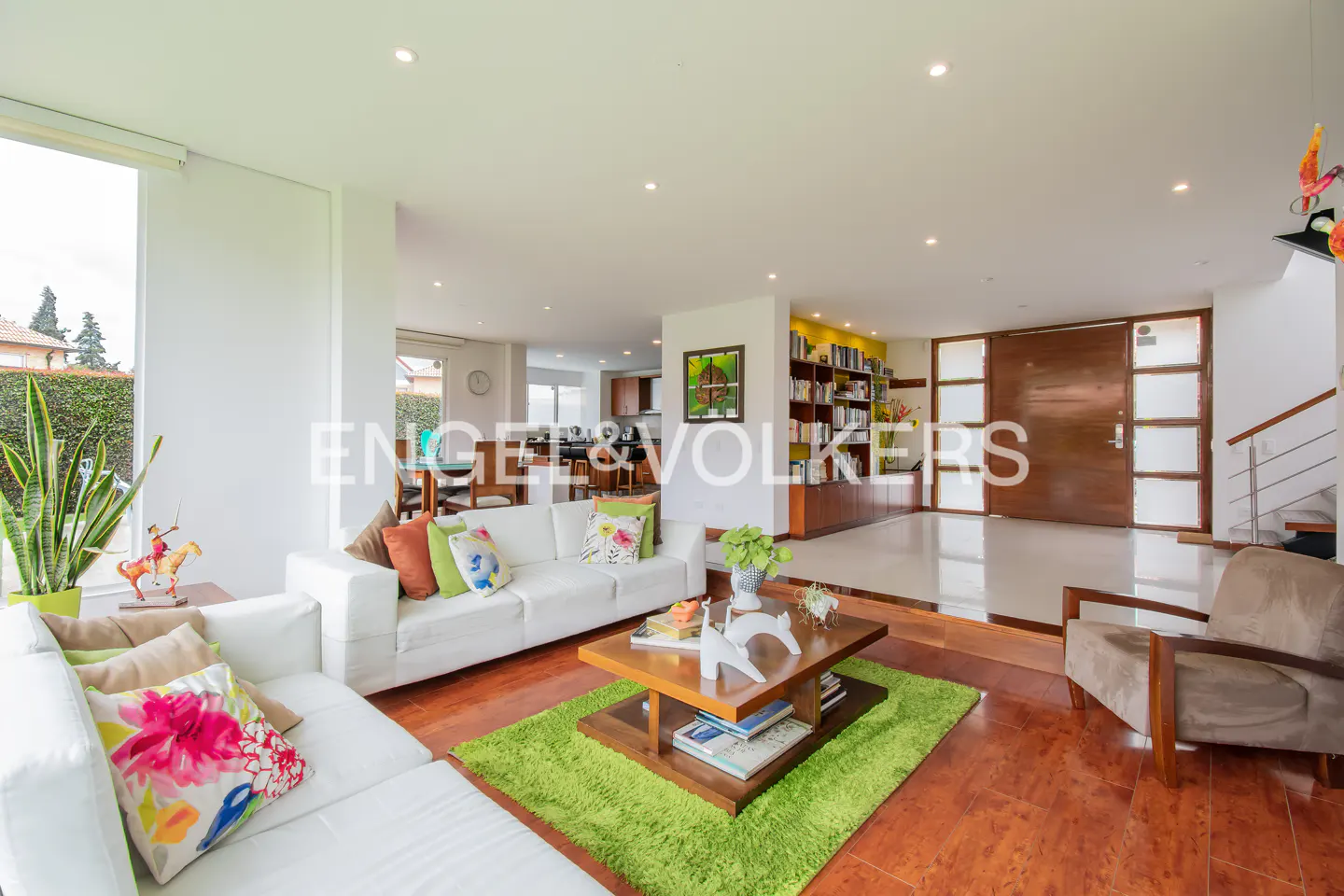 Bright living room with white sofas, colorful pillows, and a wooden coffee table on a green rug. Hardwood floors and a large window.
