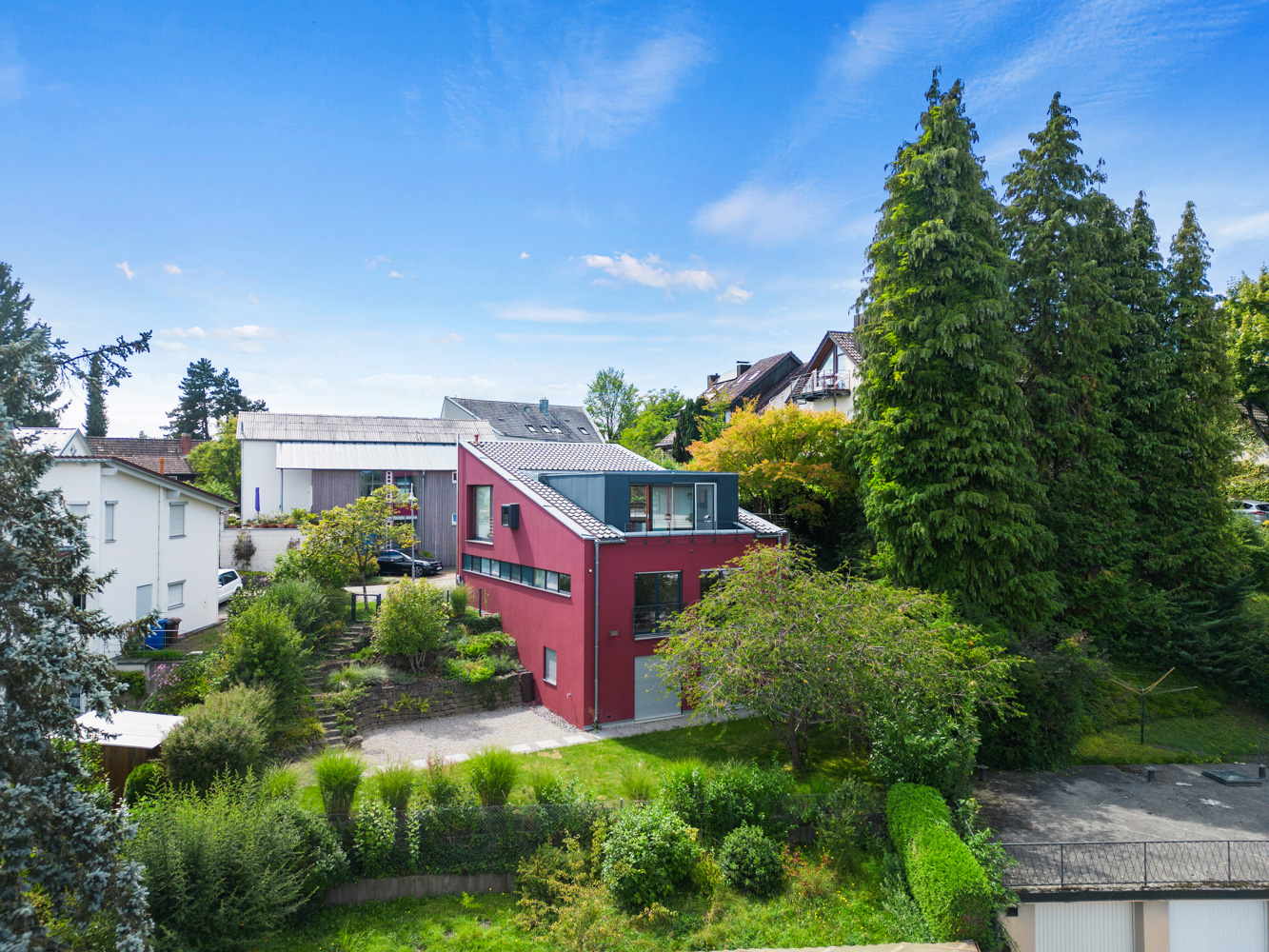 A modern, red two-story house with a slanted roof sits in a lush, green residential neighborhood under a blue sky.
