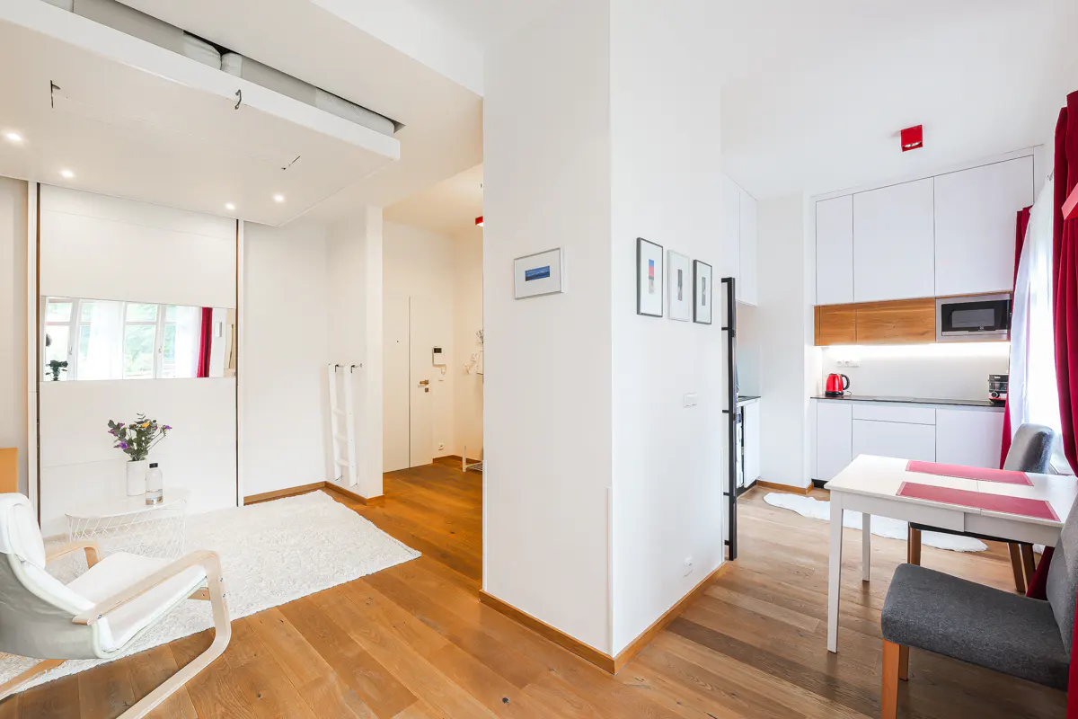 Modern studio apartment with white walls, wood floors, a white armchair, a dining area, and a sleek white kitchen.