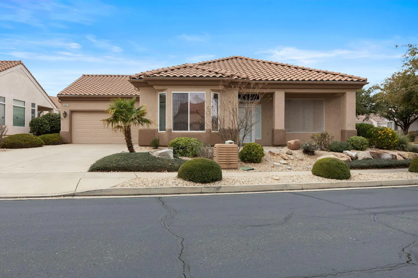 Beige single-story house with a brown tile roof, a garage, and a small palm tree in the front yard under a blue sky.