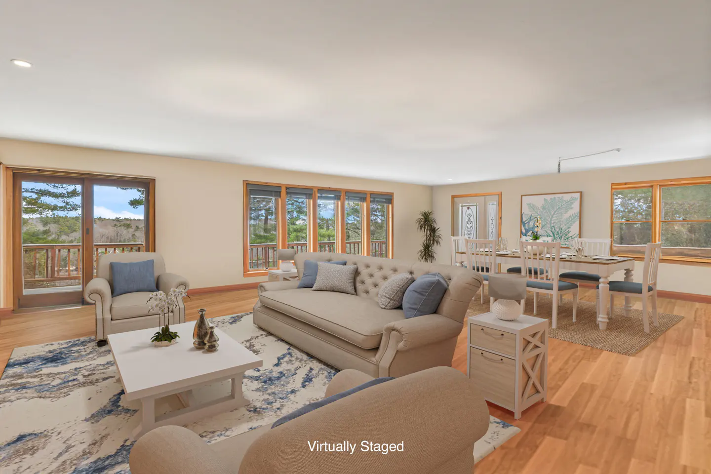 Staged living room with beige sofa, blue pillows, white table, and dining area with a nature view.
