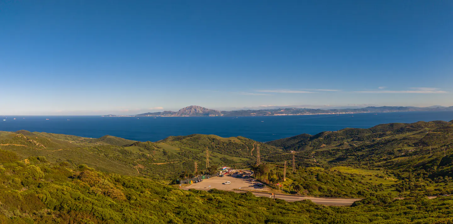 Scenic view of green hills, blue ocean, and clear sky. Distant landmass visible across the water. A parking area and road wind through the landscape.