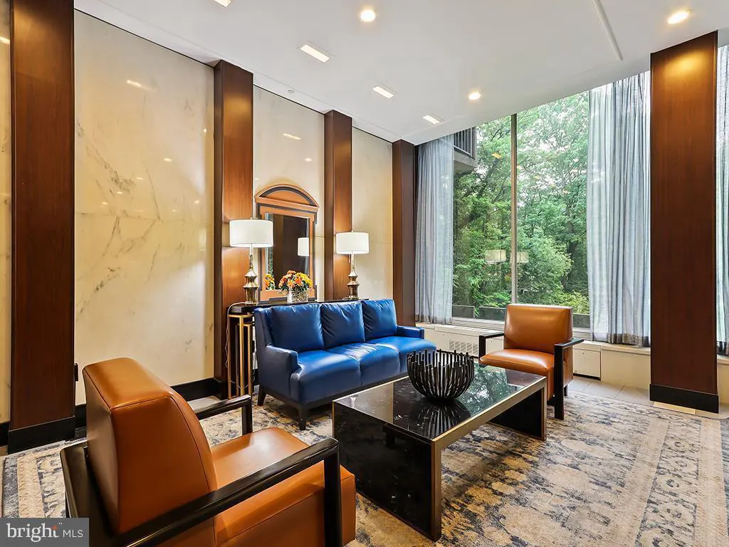 A living room with a blue sofa, two brown chairs, and a black coffee table. A large window overlooks a green forest.