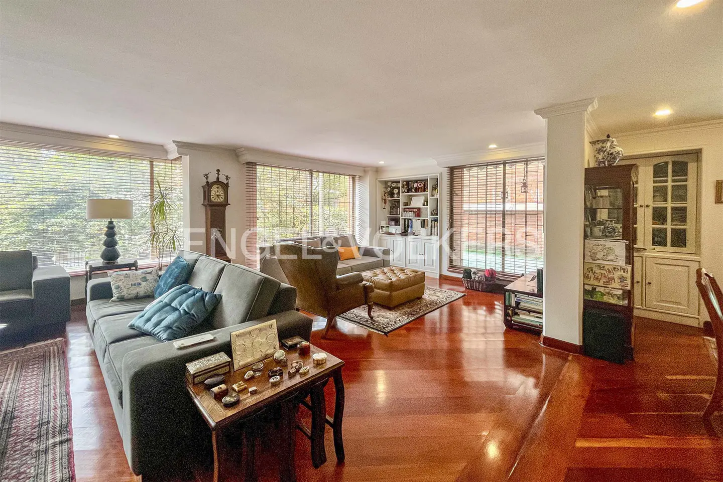A living room with hardwood floors, a gray sofa with blue pillows, and large windows with blinds.