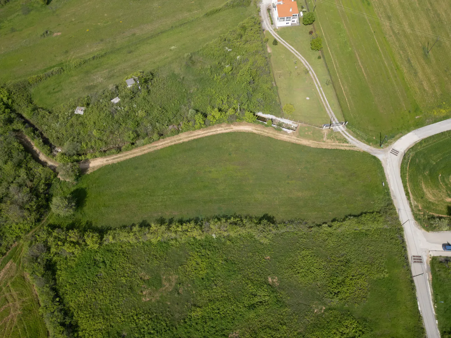 Aerial view of a green field bordered by trees and a dirt road, with a house and paved road nearby.